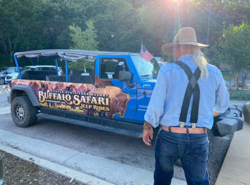 A man in a hat stands beside a blue jeep with a "Buffalo Safari Jeep Rides" sign, parked in a sunny area.