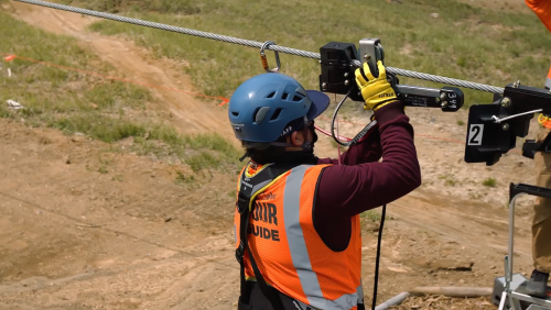 A person in a blue helmet and orange vest operates a zip line mechanism on a hillside.