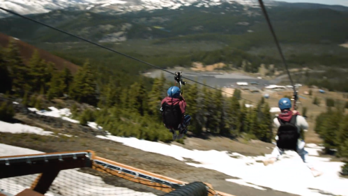 A person zips down a zip line over a snowy landscape with mountains and trees in the background.