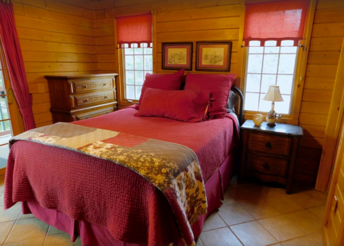 Cozy bedroom with a wooden interior, featuring a queen bed with burgundy bedding, a nightstand, and large windows.