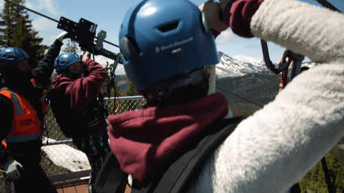 Two people in helmets prepare for a zipline ride with mountains in the background on a sunny day.