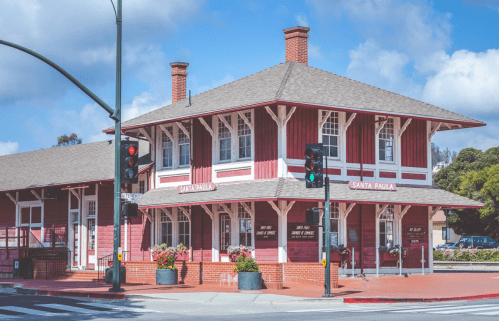Historic Santa Paula train station with red wooden exterior, surrounded by greenery and blue sky. Traffic lights visible.