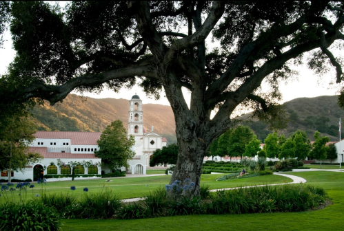 A scenic view of a campus with a large tree, buildings, and mountains in the background during sunset.