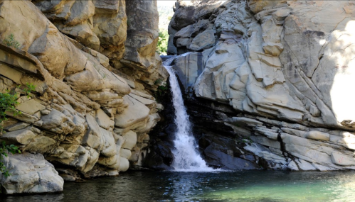 A serene waterfall cascades between rocky cliffs into a calm pool below, surrounded by lush greenery.