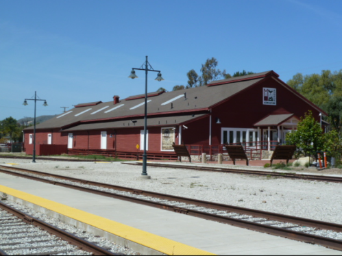 A red train station building with a sloped roof, surrounded by tracks and lampposts under a clear blue sky.