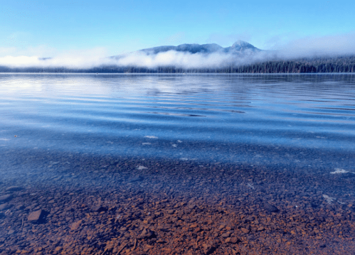 Calm lake with clear water, reflecting mountains and fog under a blue sky. Peaceful natural landscape.