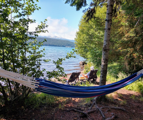 A hammock hangs between trees by a lake, with two chairs and a person relaxing in the background under a clear blue sky.