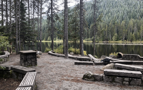 A serene lakeside scene with wooden benches, surrounded by tall trees and mountains in the background.
