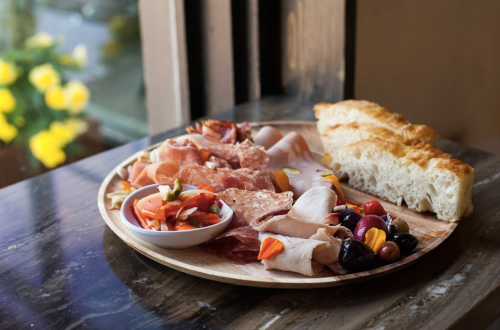 A wooden platter with assorted cured meats, olives, pickled vegetables, and a piece of bread, set near a window.