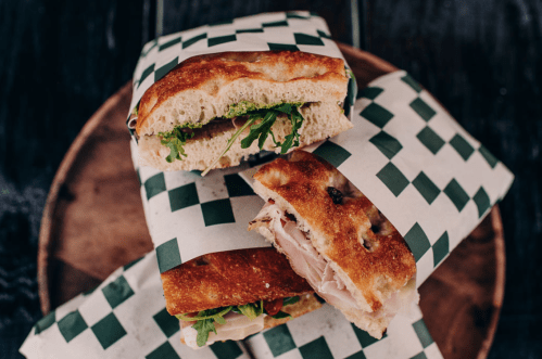 Two sandwiches wrapped in green and white checkered paper, resting on a wooden plate. One sandwich is partially visible.