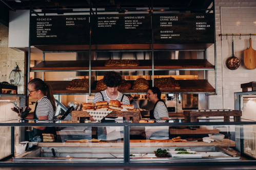 Three staff members work behind a counter in a bustling eatery, preparing food and serving customers.