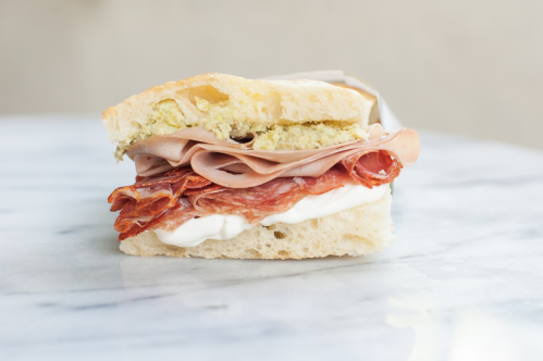 A close-up of a sandwich with layers of salami, ham, and mayonnaise on crusty bread, set on a marble surface.