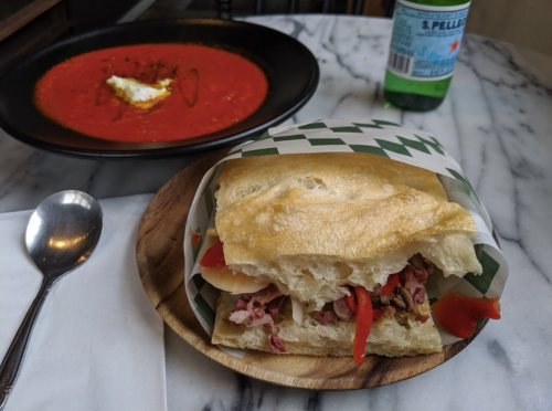A sandwich with meat and peppers on a wooden plate, alongside a bowl of red soup and a sparkling water bottle.
