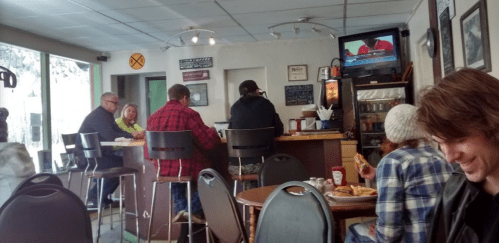 A cozy café interior with patrons seated at tables and a counter, watching a TV in the background.