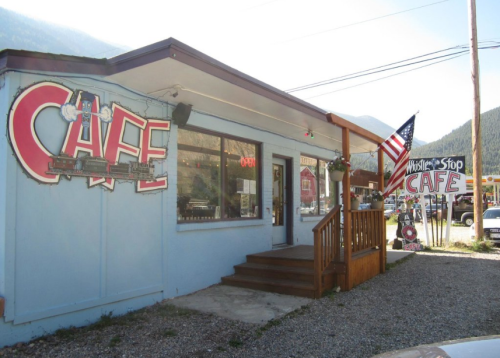 A quaint cafe with a blue exterior, large "CAFE" sign, and an American flag, set in a mountainous area.
