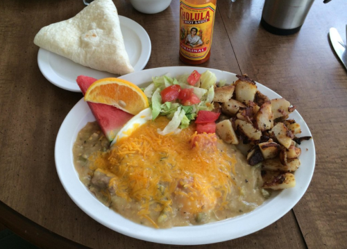 A plate of hearty breakfast featuring eggs, cheese, potatoes, salad, watermelon, orange, and a tortilla, with hot sauce.
