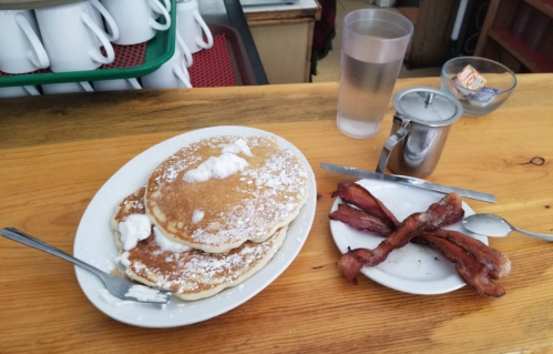 A plate of fluffy pancakes topped with powdered sugar and whipped cream, alongside a side of crispy bacon.