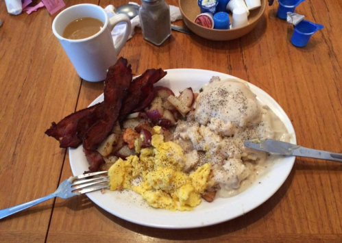 A plate of breakfast with scrambled eggs, bacon, potatoes, biscuits with gravy, and a cup of coffee on a wooden table.