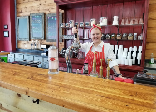 A smiling employee in a retro-style diner serves drinks from a soda fountain at a wooden counter.
