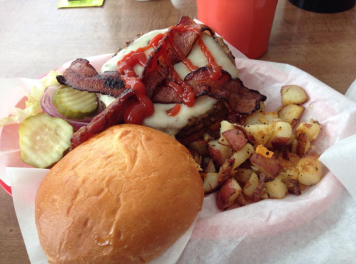 A hearty burger topped with cheese, bacon, and ketchup, served with pickles, onions, and seasoned potato wedges.