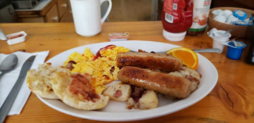 A plate of breakfast featuring scrambled eggs, sausage links, potatoes, a biscuit, and an orange slice.