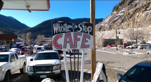 Sign for "Whistle Stop Cafe" in a snowy mountain town, with parked cars and rocky hills in the background.