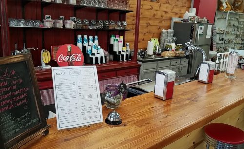 A cozy café counter with a menu, Coca-Cola sign, and various drink dispensers in a rustic setting.
