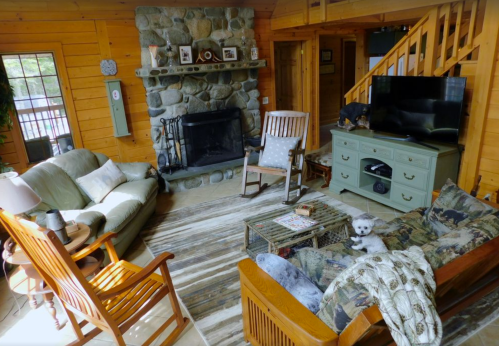 Cozy living room with a stone fireplace, wooden furniture, and a small dog on a couch. Warm, rustic decor.