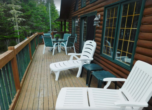 A wooden deck with white lounge chairs and a table, surrounded by trees and a cozy cabin.