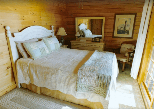 Cozy bedroom with a wooden interior, featuring a white bed, dresser, mirror, and a chair by the window.