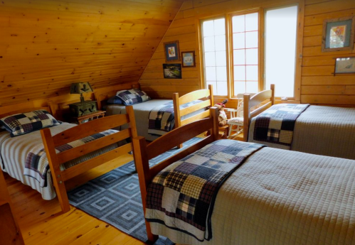Cozy attic bedroom with wooden walls, featuring four twin beds, quilts, and large windows for natural light.