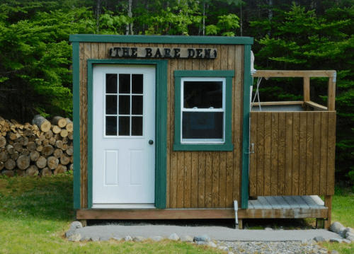 A small wooden cabin named "The Bare Den," featuring a white door and a porch, surrounded by trees and firewood.