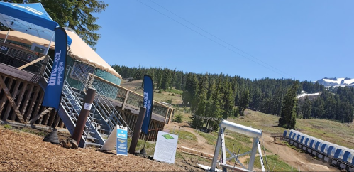 A ski resort area with a wooden deck, flags, and a view of green hills and snow-capped mountains in the background.