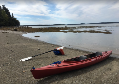 A red kayak rests on a sandy shore beside calm water, with a paddle and backpack nearby under a cloudy sky.