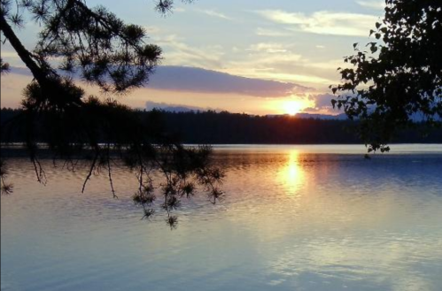 A serene sunset over a calm lake, with trees framing the scene and colorful clouds reflecting on the water.