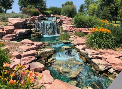 A serene waterfall cascades over pink rocks into a clear pool, surrounded by vibrant flowers and lush greenery.