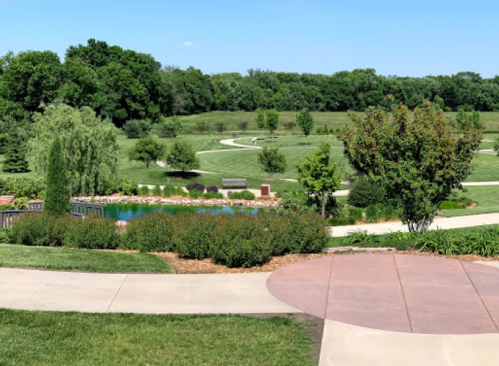 A scenic park with winding paths, lush greenery, and a small pond surrounded by trees and shrubs under a clear blue sky.