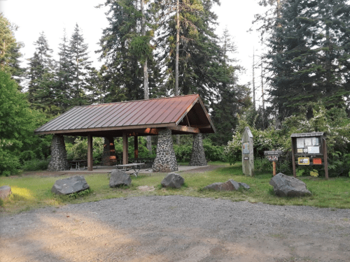 A rustic picnic shelter with a metal roof, surrounded by trees and rocks, in a grassy area. Informational signs nearby.