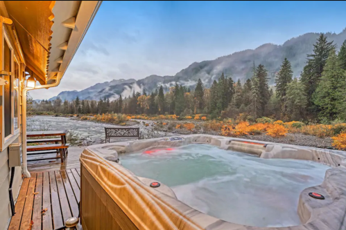 A hot tub on a wooden deck overlooks a misty river and mountains, surrounded by autumn foliage.