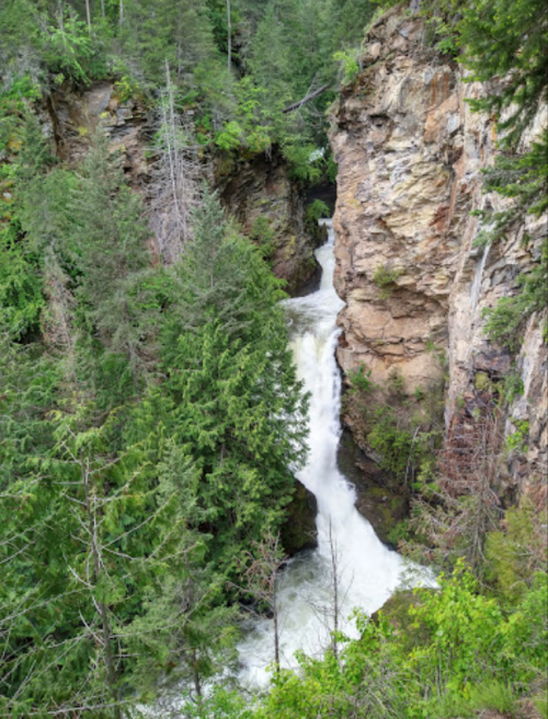 A waterfall cascades through a narrow canyon surrounded by lush green trees and rocky cliffs.