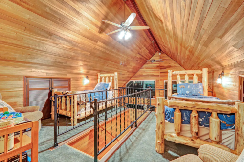 Cozy loft bedroom with wooden walls, two log-style beds, and a ceiling fan, overlooking a balcony.