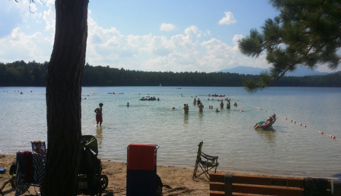 People swimming and playing in a lake on a sunny day, with trees and mountains in the background.