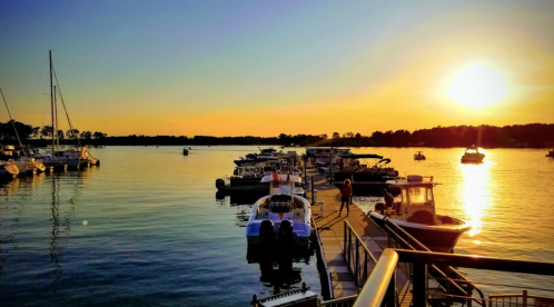 Sunset over a marina with boats docked and people walking along the pier, reflecting golden hues on the water.