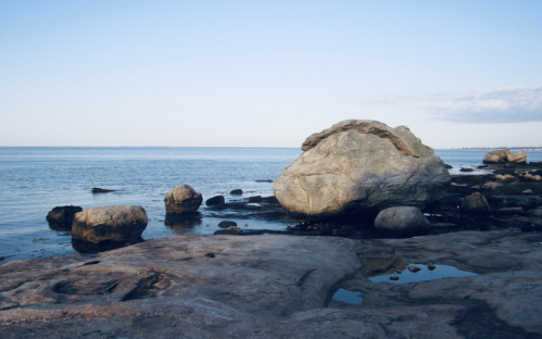 A serene coastal scene featuring large rocks on a sandy shore with calm water and a clear blue sky.