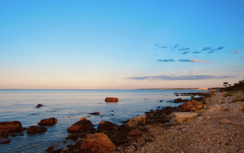 A serene beach scene at sunset, featuring calm waters, rocky shoreline, and a clear blue sky with scattered clouds.