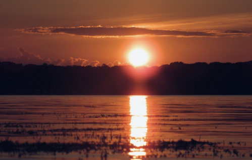 A vibrant sunset over a calm lake, with reflections shimmering on the water and silhouettes of trees in the background.