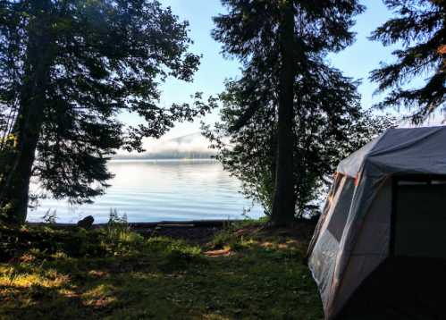 A serene lakeside view with a tent, surrounded by trees and morning mist over the water.