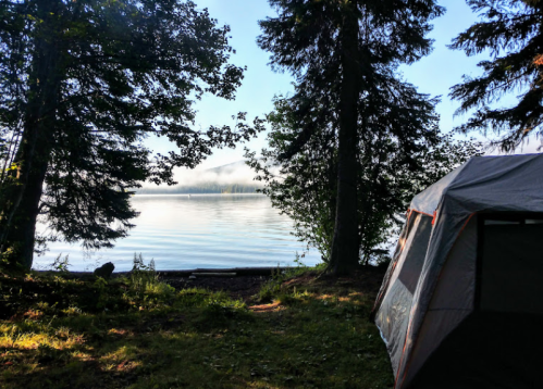 A serene lakeside view with a tent, surrounded by trees and morning mist over the water.