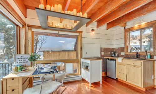 Cozy kitchen with wooden beams, modern appliances, and a dining table set for two, featuring large windows and natural light.