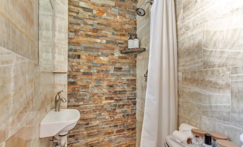 A modern shower with a stone wall, white curtain, sink, and neatly arranged toiletries.